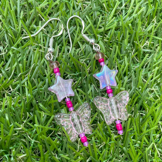 Butterfly-shaped earrings with star charms and pink beads on a grass background