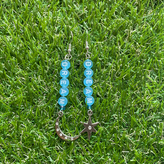 Pair of earrings with blue circular beads spelling "dream" and ending in a moon and star lying on grass